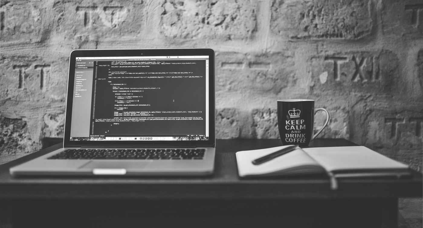 Grayscale photo of MacBook, disolaying code editor, near white notebook and ceramic mug on table